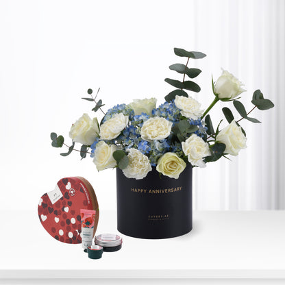 Elegant black anniversary flower arrangement with white roses and blue hydrangeas, accompanied by a heart-shaped chocolate box and skincare set.