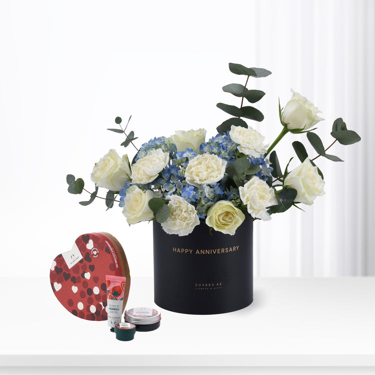 Elegant black anniversary flower arrangement with white roses and blue hydrangeas, accompanied by a heart-shaped chocolate box and skincare set.