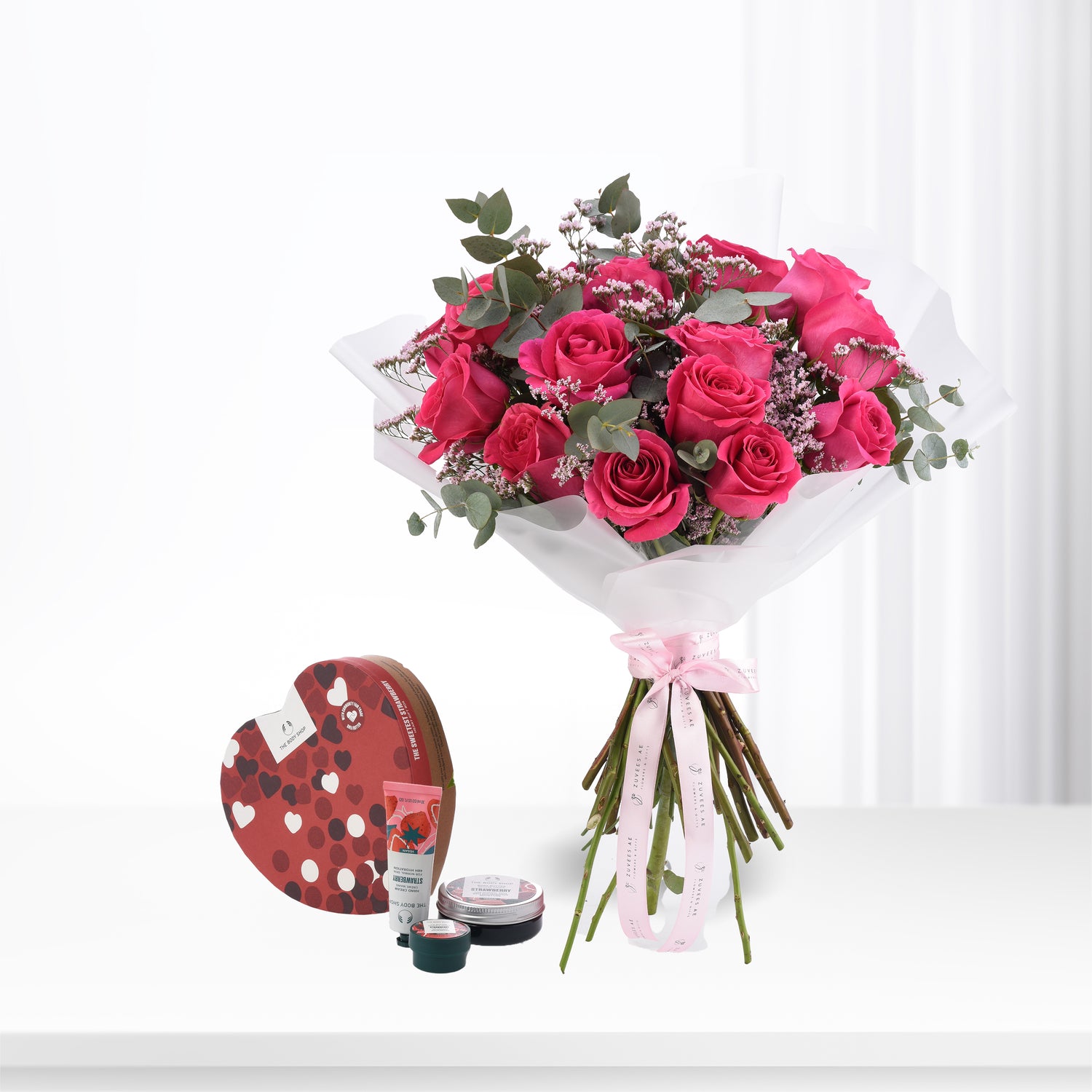 Vibrant pink roses with eucalyptus, wrapped in white paper and pink ribbon, paired with a heart-shaped The Body Shop gift set.
