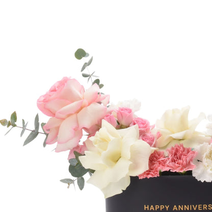 Soft pink and cream roses with eucalyptus in black box, "Happy Anniversary" gold text.