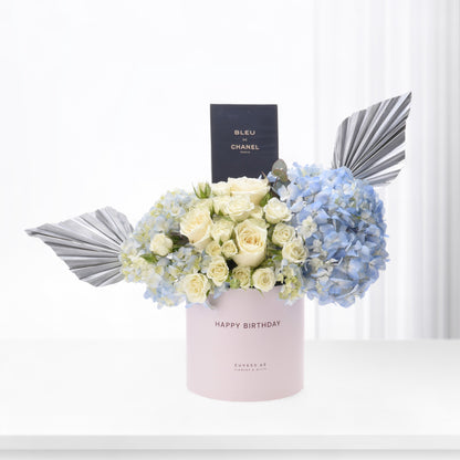 Pastel pink box holds creamy roses and blue hydrangeas, accented with silver fan leaves and a Chanel perfume box.