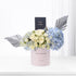 Pastel pink box holds creamy roses and blue hydrangeas, accented with silver fan leaves and a Chanel perfume box.