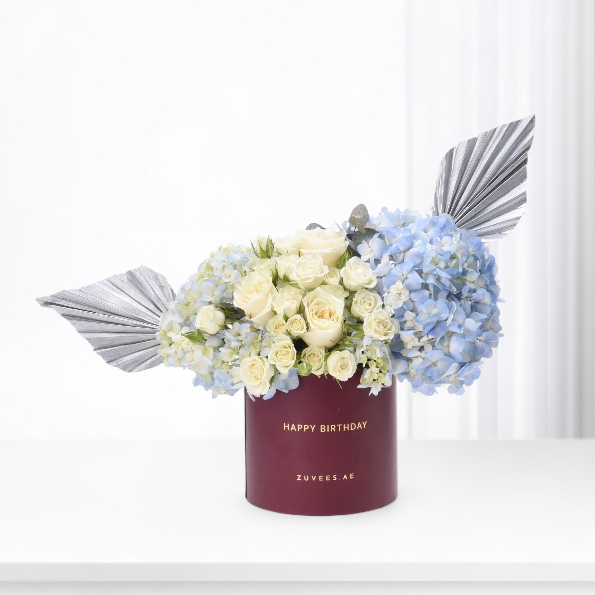 Elegant white roses, blue hydrangeas, silver foliage in burgundy "Happy Birthday" box, minimalist luxury floral gift.