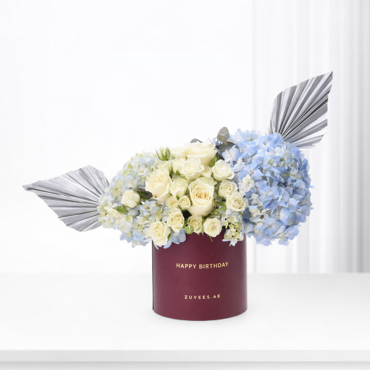Elegant white roses, blue hydrangeas, silver foliage in burgundy "Happy Birthday" box, minimalist luxury floral gift.