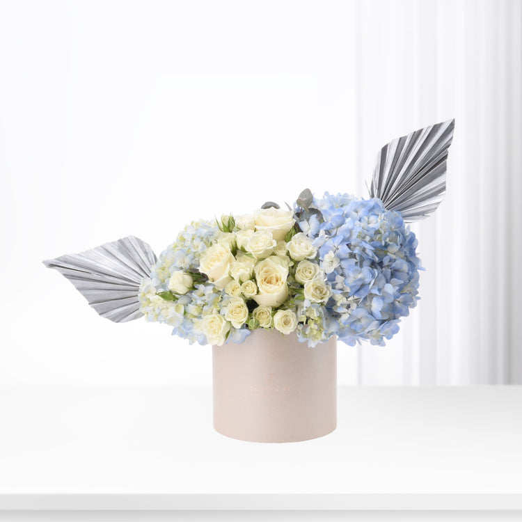 Soft blue hydrangeas and cream roses in blush box with silver fan accents.