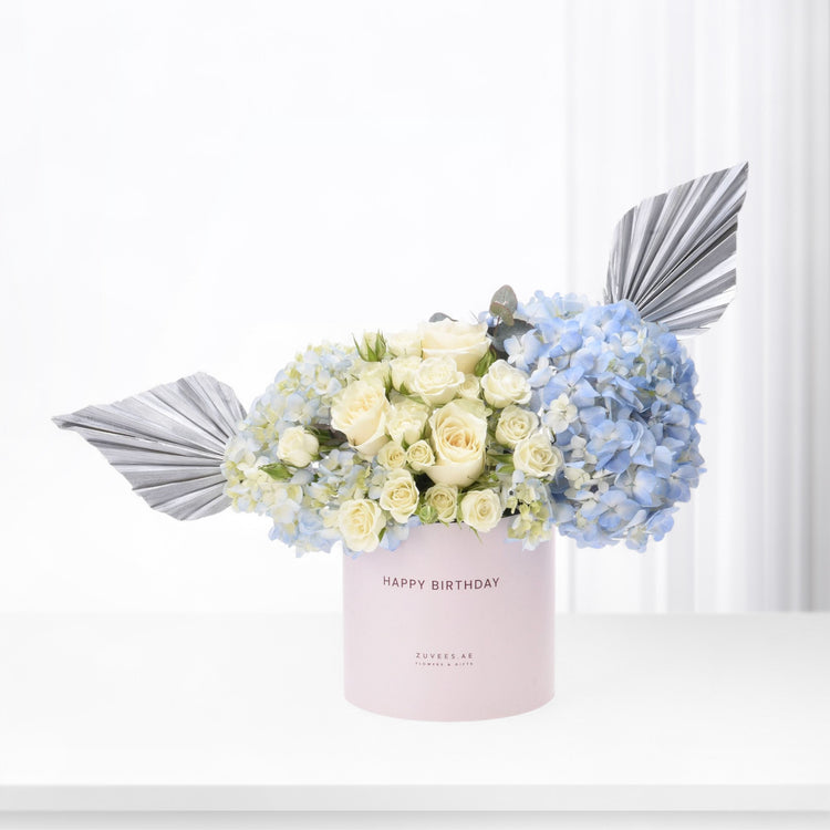 Pastel pink box holds creamy roses and blue hydrangeas, accented with silver fan leaves.
