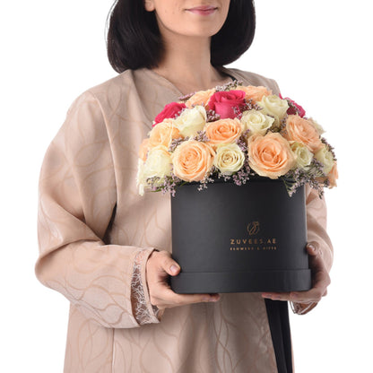 Elegant black box holds vibrant peach, cream, and pink roses with lavender accents.