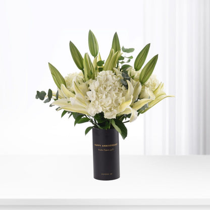 Elegant white lilies, hydrangeas in sleek black "Happy Anniversary" vase, minimalist modern design.