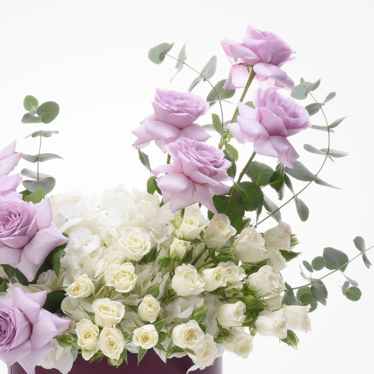 Delicate lavender roses, creamy white blooms, and eucalyptus in elegant purple vase.