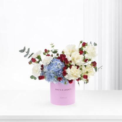 Pink cylindrical vase holds white roses, red buds, and blue hydrangeas against bright white background.