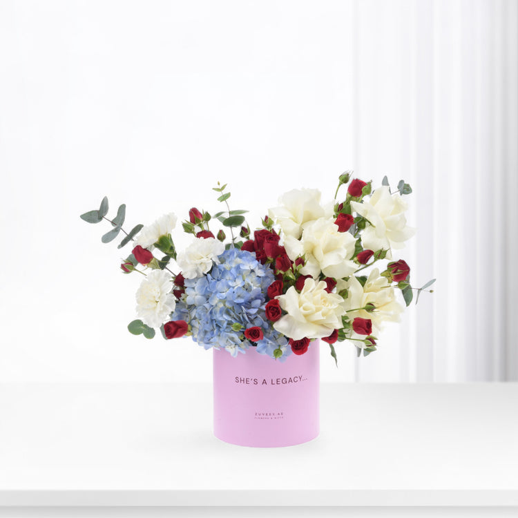 Pink cylindrical vase holds white roses, red buds, and blue hydrangeas against bright white background.
