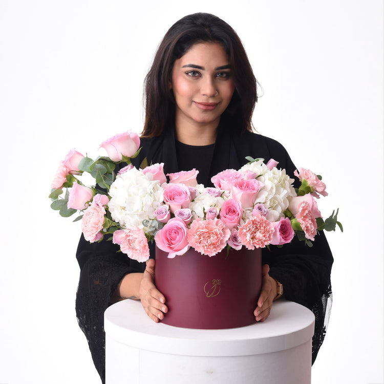 Elegant woman holds luxurious burgundy floral box overflowing with pink roses, carnations, and white hydrangeas.