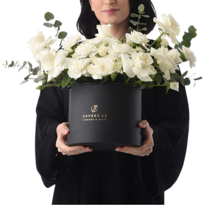 Elegant white roses and eucalyptus in sleek black Zuvees box, held by woman in black.