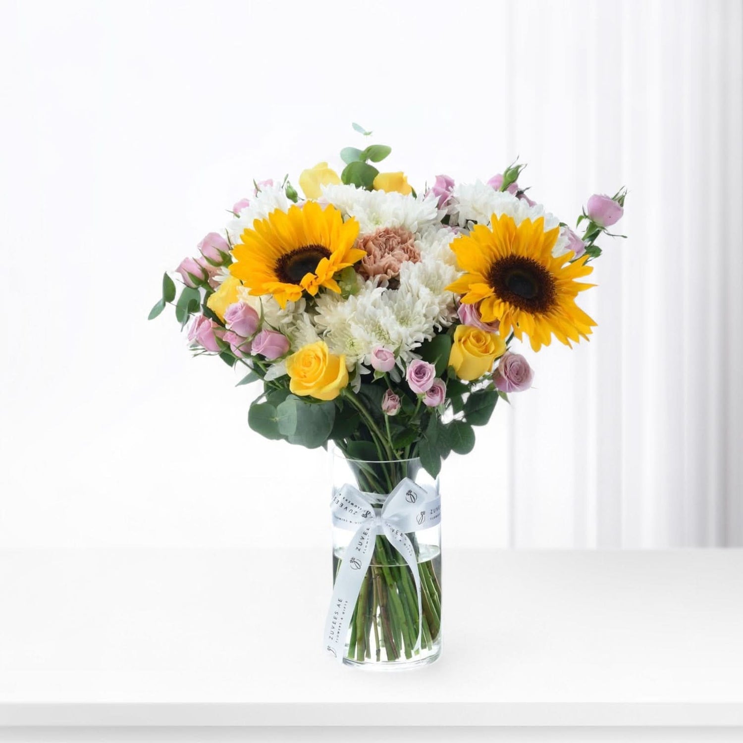 Vibrant sunflowers, roses, and chrysanthemums in clear vase with branded bow.