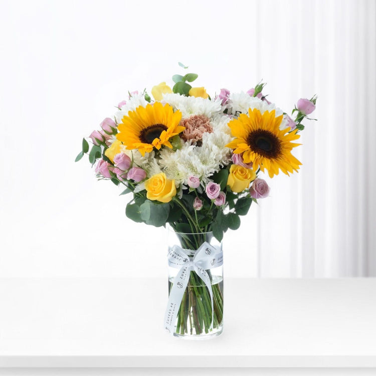 Vibrant sunflowers, roses, and chrysanthemums in clear vase with branded bow.