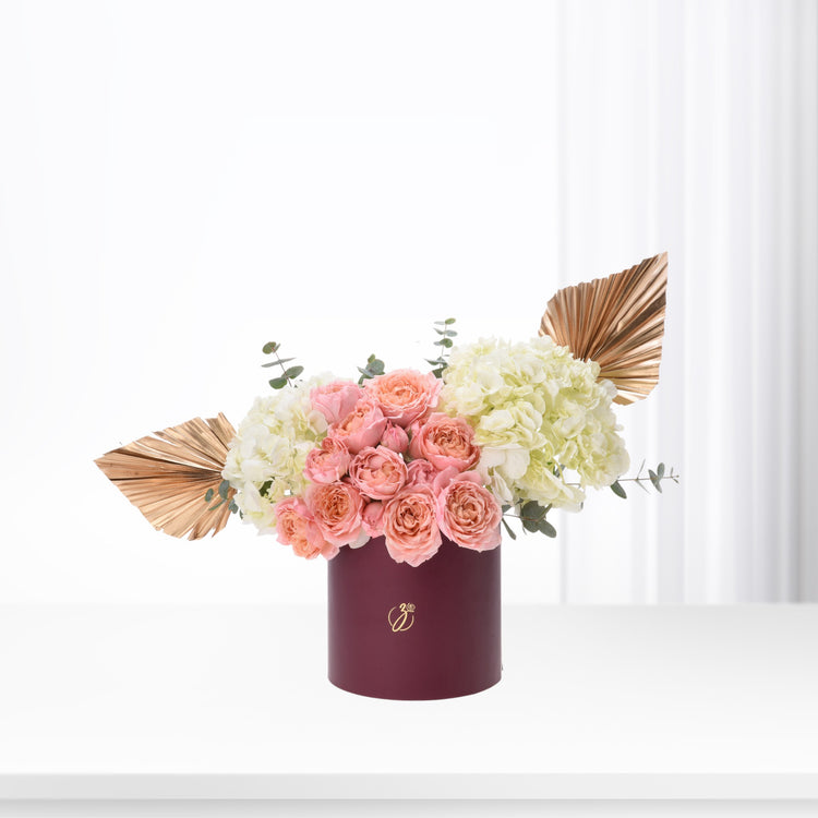 Elegant burgundy box holds blush roses and cream hydrangeas, accented with gold fan leaves.
