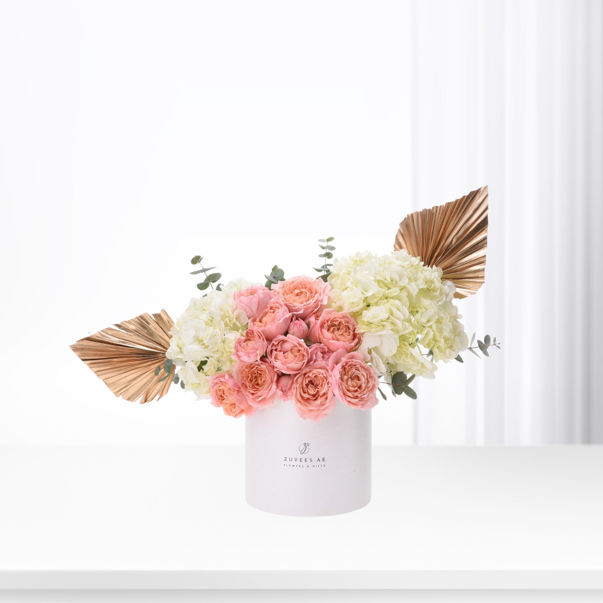 Elegant pink roses, white hydrangeas, gold palm leaves in white Zuvees box, minimalist luxury floral arrangement.