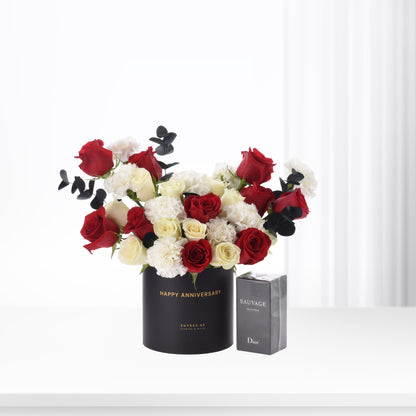 Elegant red, white roses and carnations in black "Happy Anniversary" vase beside Dior Sauvage perfume.