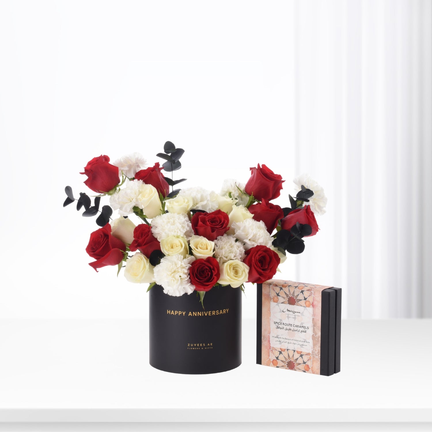 Elegant red and white roses with black eucalyptus in black "Happy Anniversary" vase, beside spice caramel gift box.