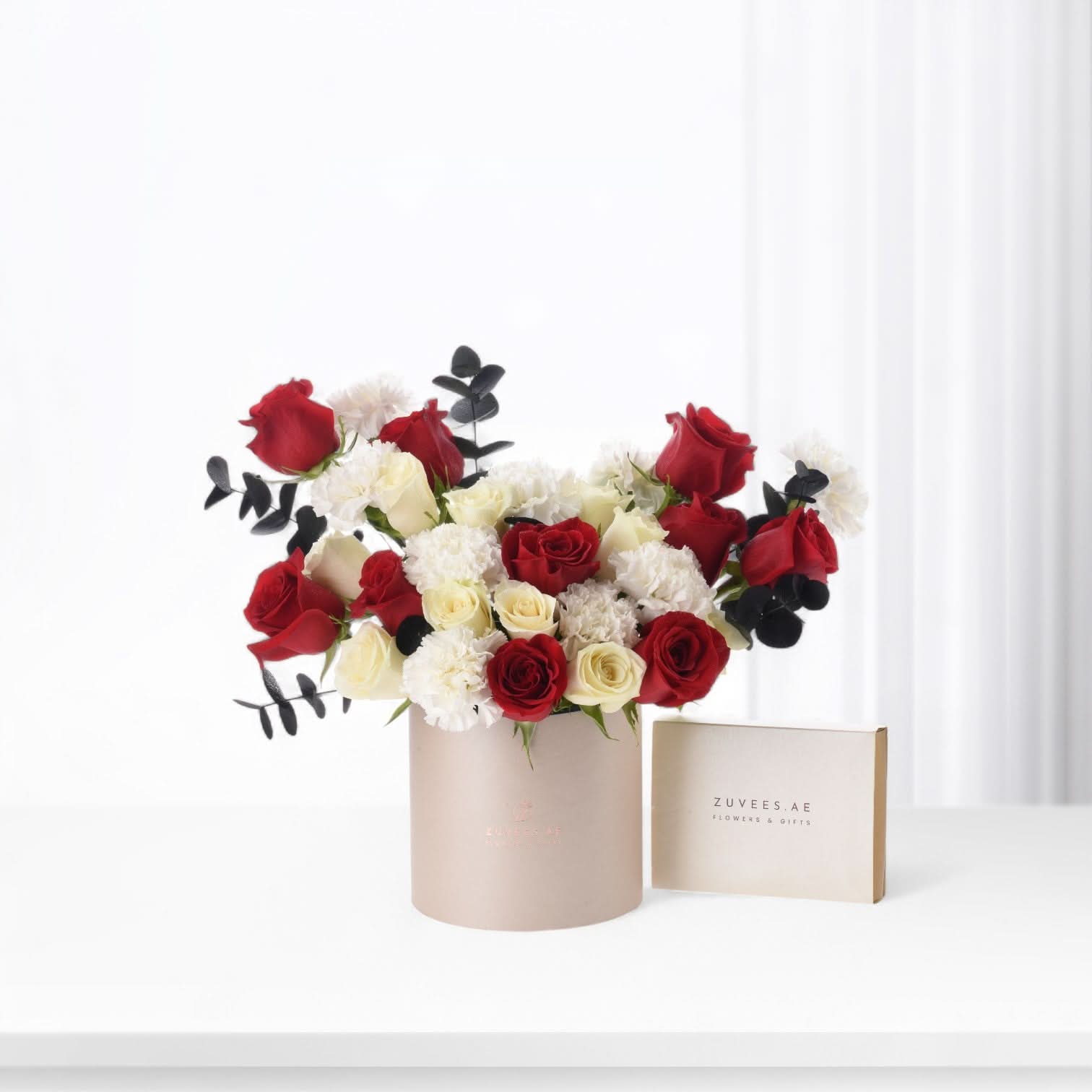 Elegant sand box arrangement with red and white roses from ZUVEES, paired with Luxe Chocolate, Mirzam, and Lakrids — a refined gift of sweetness and love.