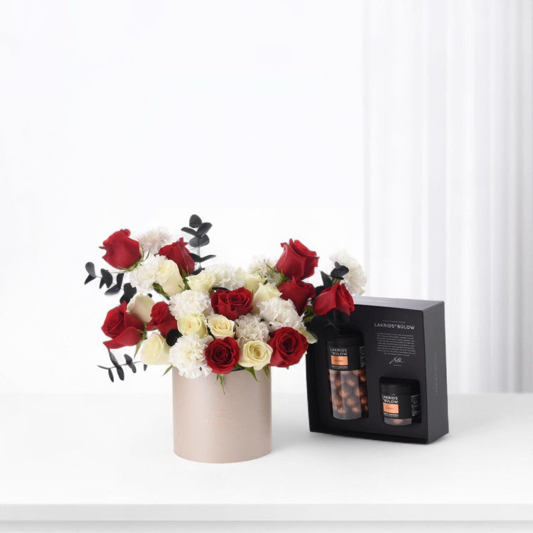 Elegant sand box arrangement with red and white roses from ZUVEES, paired with Luxe Chocolate, Mirzam, and Lakrids — a refined gift of sweetness and love.