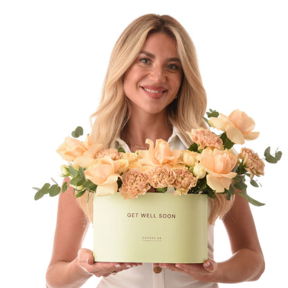 Smiling woman holds pastel peach floral box reading "Get Well Soon".