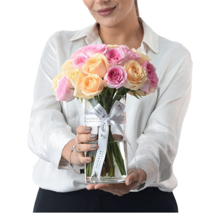 Woman holds clear vase with pastel pink and peach roses, branded ribbon.