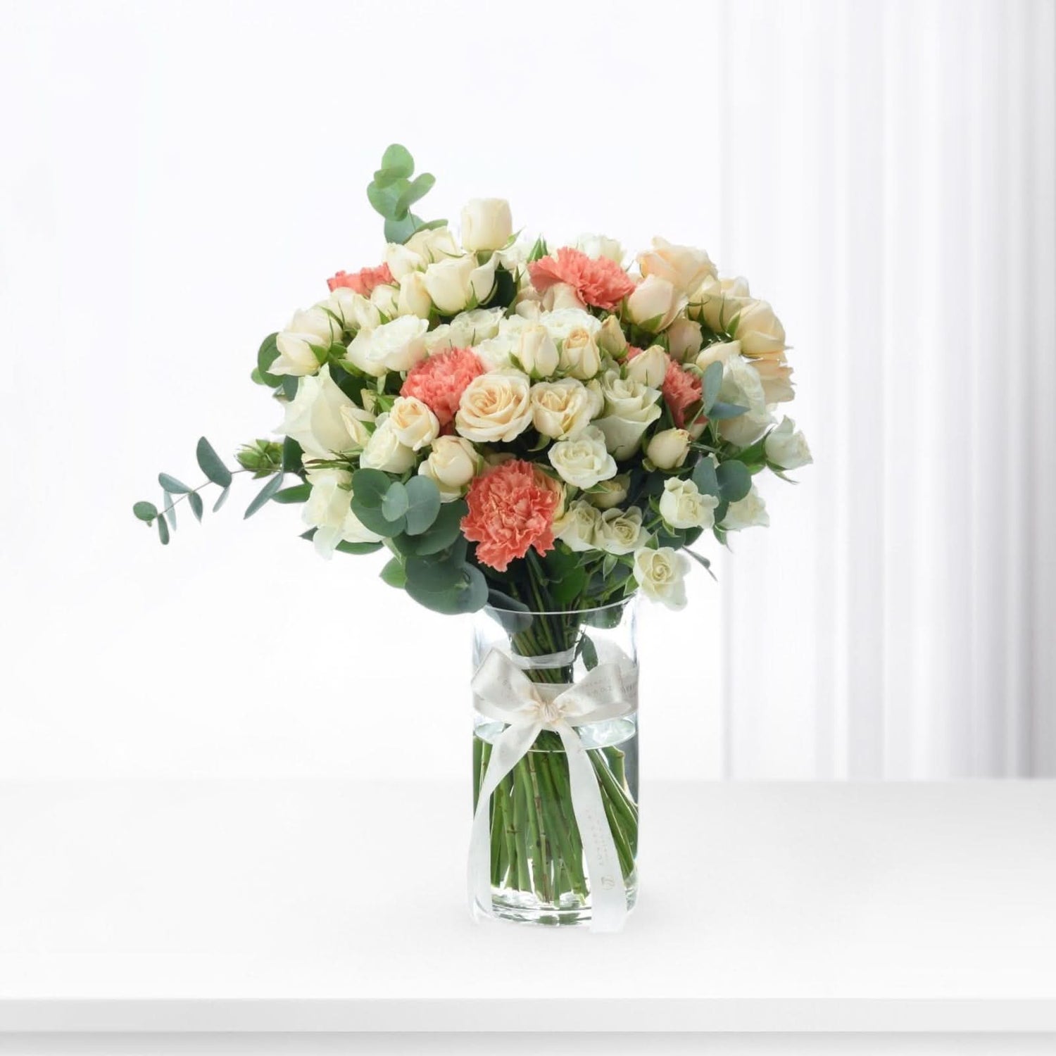 White roses and coral carnations in clear vase with silk bow.