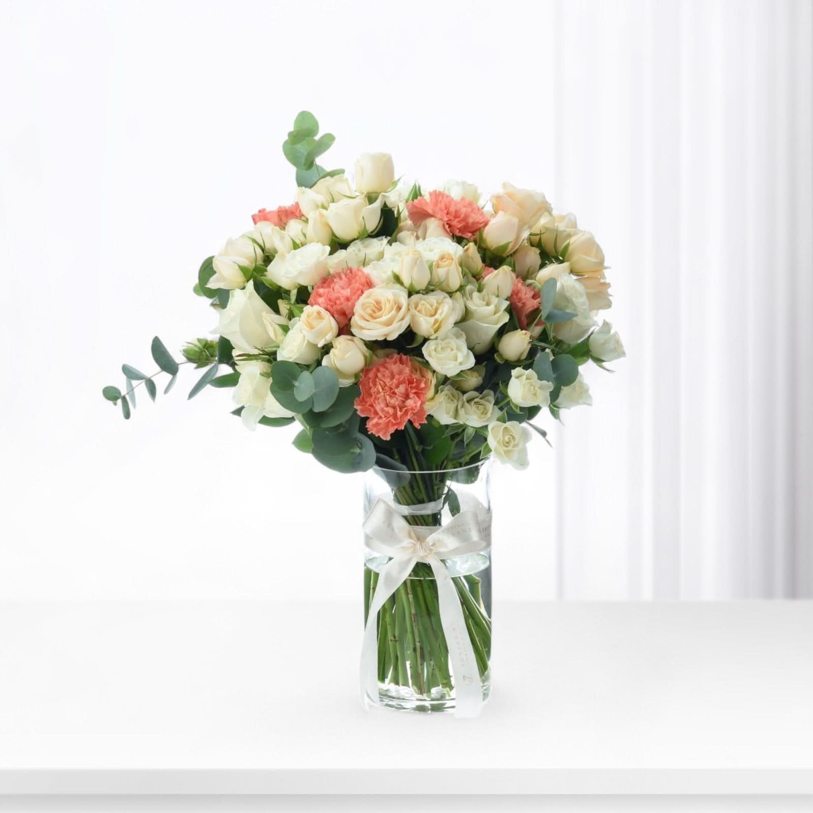 White roses and coral carnations in clear vase with silk bow.