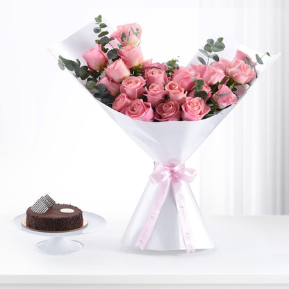Pink roses in white wrap with pink ribbon, beside chocolate cake on white stand.