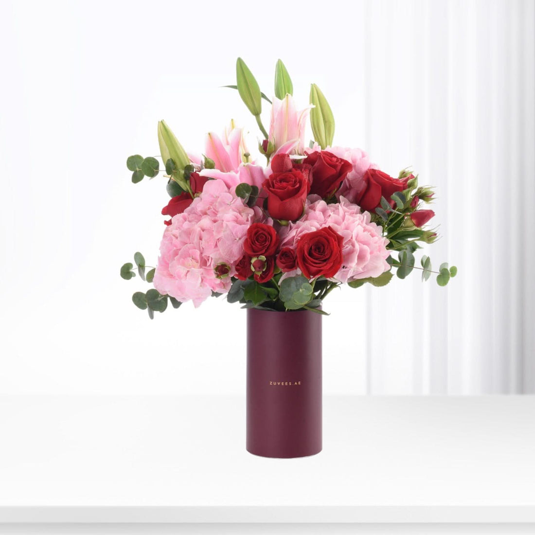Vibrant red roses, pink hydrangeas, and lilies in deep burgundy vase, elegant floral gift.