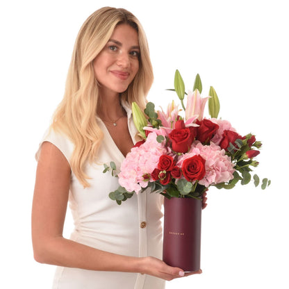 Woman holds elegant bouquet of red roses, pink hydrangeas, and lilies in matte burgundy vase.