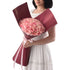 Woman holds lush pink rose bouquet in rich burgundy wrap with delicate pink ribbon.