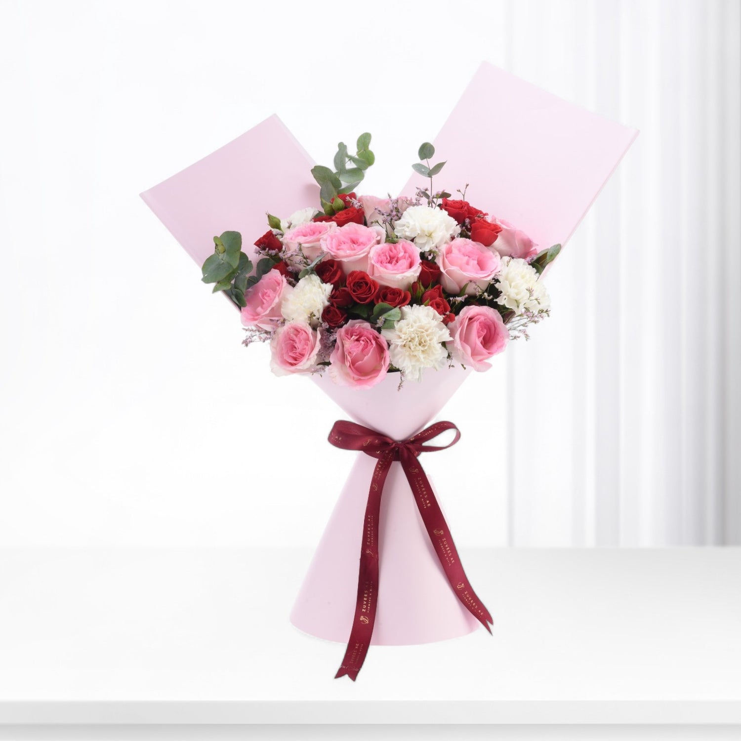 Pink-wrapped bouquet with red, pink, and white roses, accented by greenery and a burgundy ribbon.