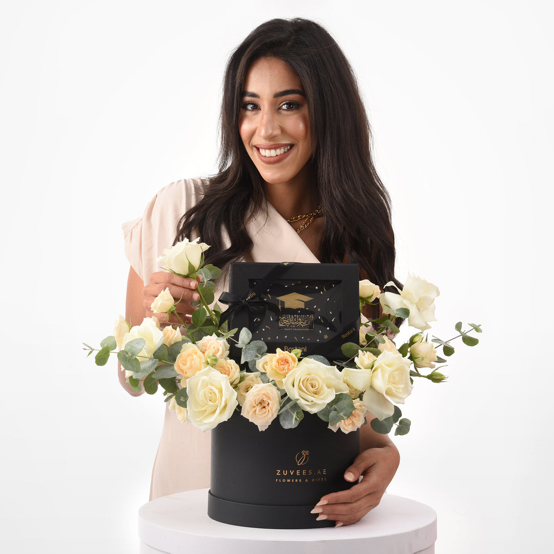 Elegant woman smiles holding black graduation-themed floral gift box with cream roses.