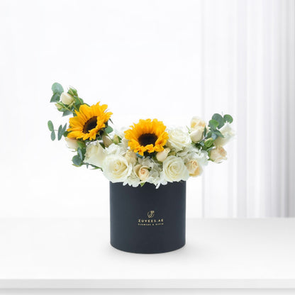 Vibrant sunflowers and white roses in sleek black box with gold logo.
