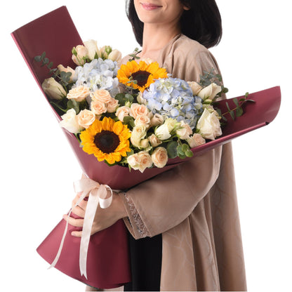 Vibrant sunflowers, roses, and hydrangeas in burgundy wrap with cream ribbon.