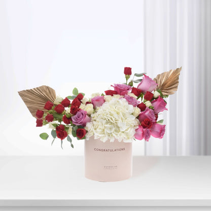 Pink "Congratulations" vase with red, pink, white roses, hydrangeas, gold palm leaves, elegant gift arrangement.