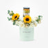 Pastel green cylinder holds sunflowers, white roses, and marmalade jar.