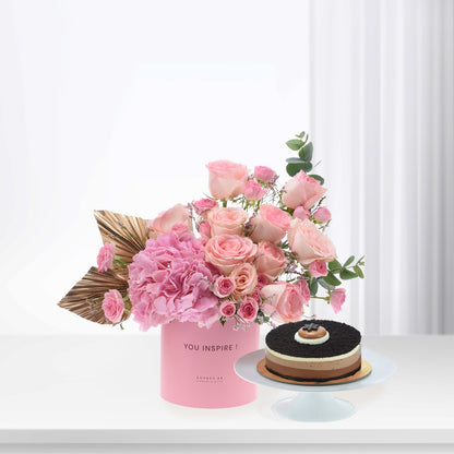 Pink roses and hydrangeas in branded box beside layered chocolate cake.