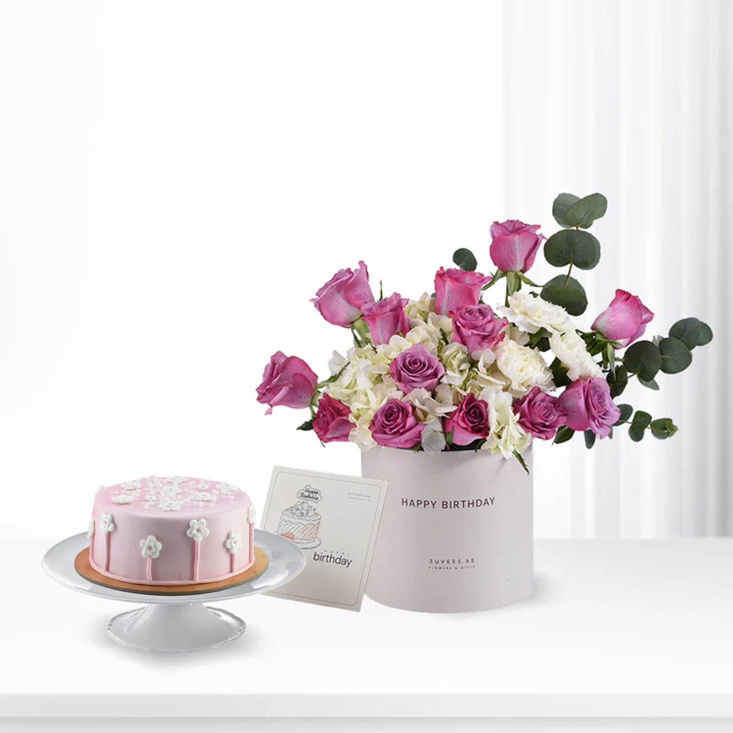 Pink cake with white flowers beside pink and white rose bouquet in "Happy Birthday" box.