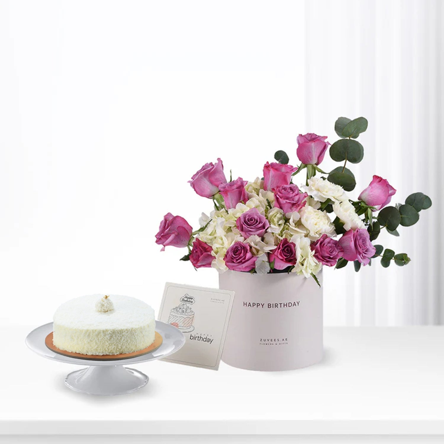 Pink roses and white hydrangeas in a "Happy Birthday" box beside a coconut cake.