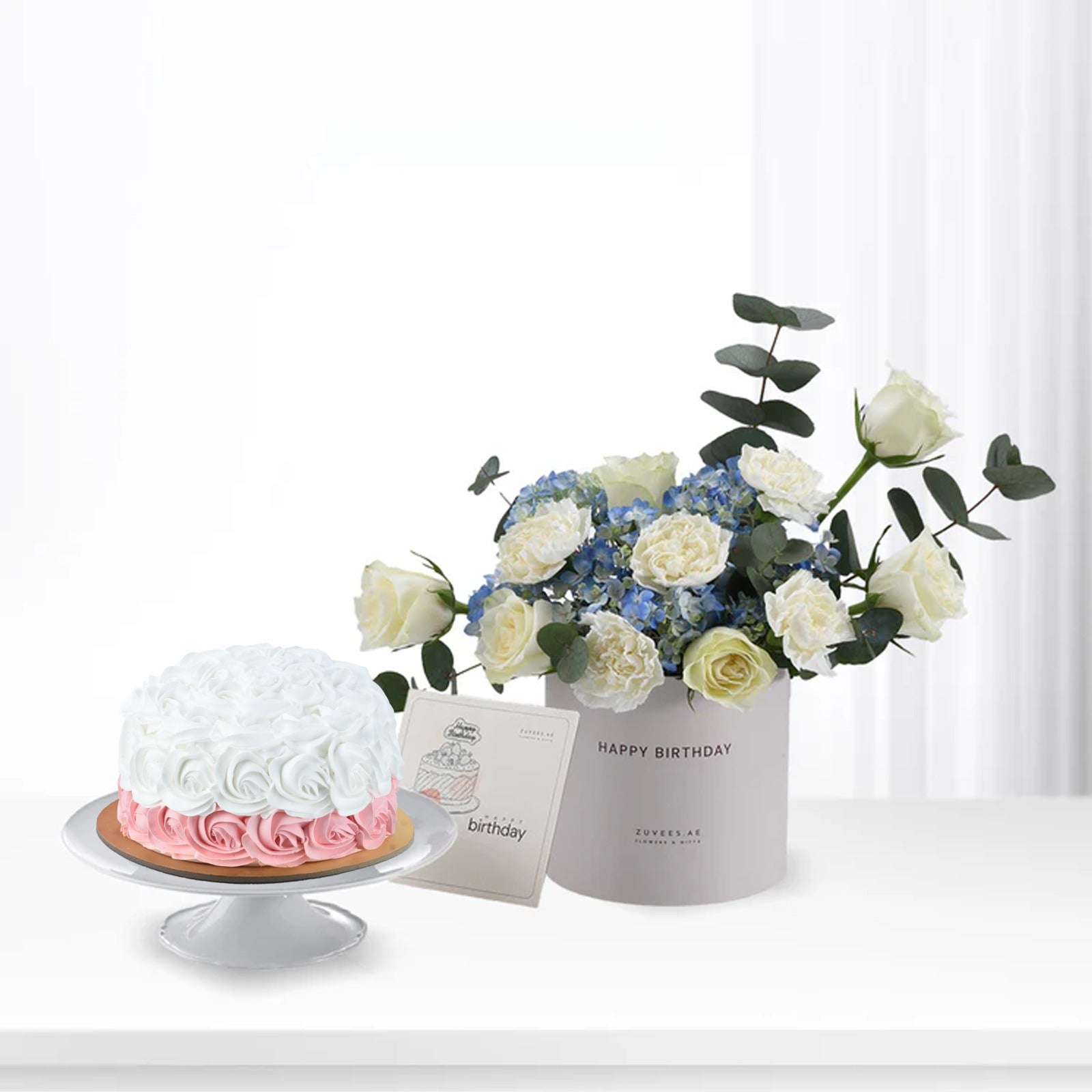 White and pink rose-patterned cake beside elegant white rose and blue hydrangea bouquet in "Happy Birthday" box.