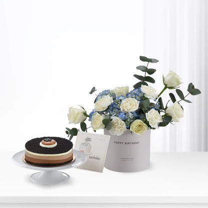 Elegant birthday gift set: chocolate cake, white roses, blue hydrangeas, and "Happy Birthday" card.