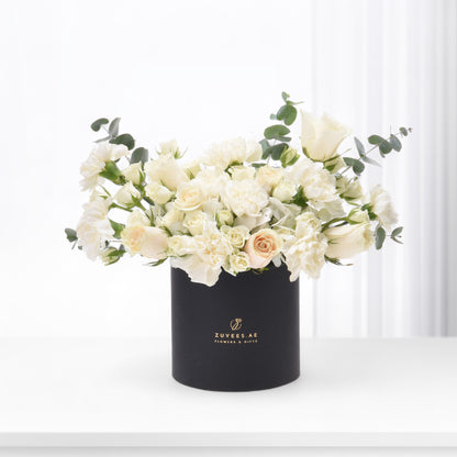 Elegant white roses and eucalyptus in matte black cylindrical vase with gold branding.