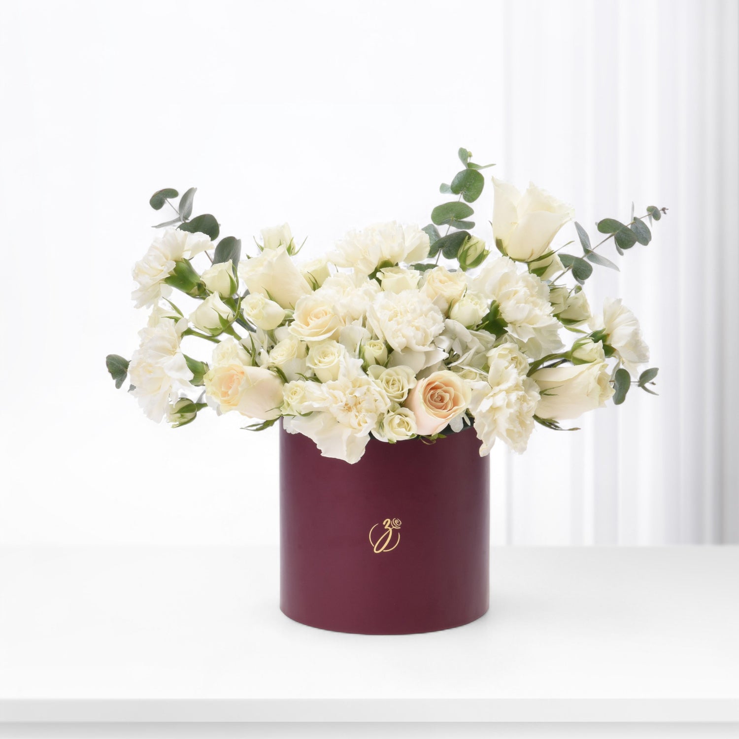 Elegant white roses and carnations in deep burgundy box with gold logo, accented by green eucalyptus.