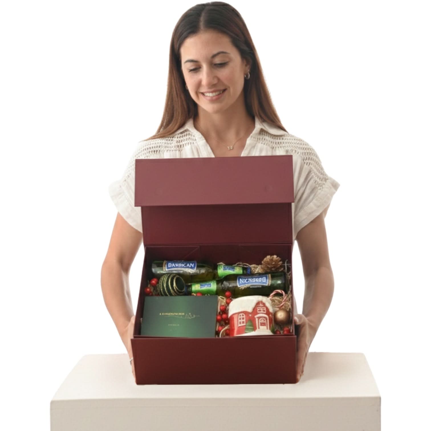 Woman holds festive red gift box with wine, ornaments, and holiday mug inside.