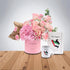 Pink roses and hydrangeas in branded pink vase, matching mug and notebook on wooden table.