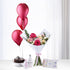 Shimmering red balloons rise beside floral bouquet, baby essentials, and chocolate cake.
