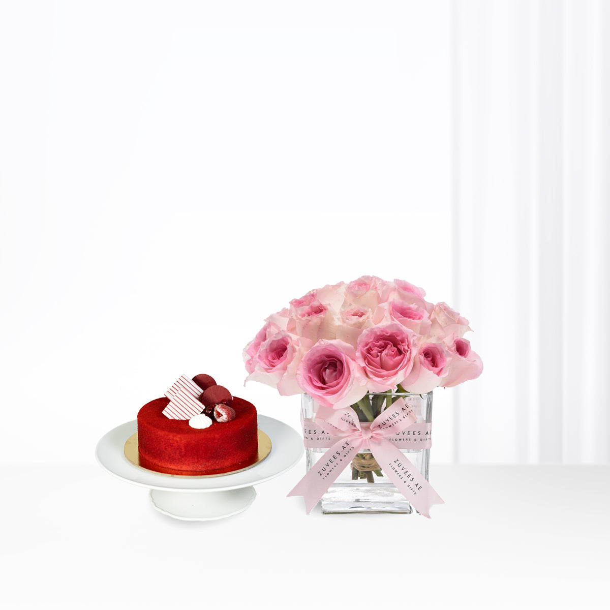 Vibrant red velvet cake beside blush pink roses in glass vase, elegant gift set.